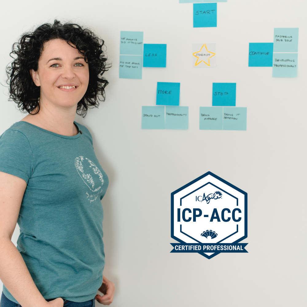 Paulina Hornbachner is standing in front of a wall with Post Its sticking on it. The Badge says "Certified Professional in Agile Coaching"