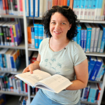 Paulina Hornbachner is standing in a library with a book in her hands and smiling in the camera.