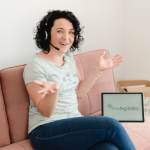 Paulina Hornbachner is sitting on a couch and speaking into her headset. The Logo "The Agilista" is visible on the Laptop's screen.